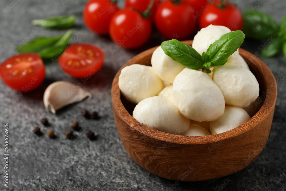 Delicious mozzarella balls in wooden bowl with basil leaves and tomatoes on light gray table, closeup. Space for text
