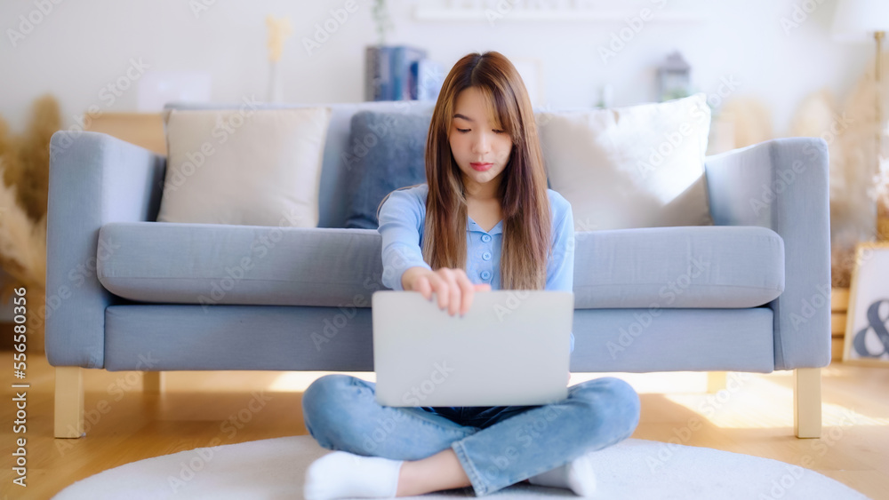 Young asian woman in good spirits working on laptop at home while sitting on the floor close to the couch Young asian woman in good spirits working on laptop at home while sitting on the floor close to the couch