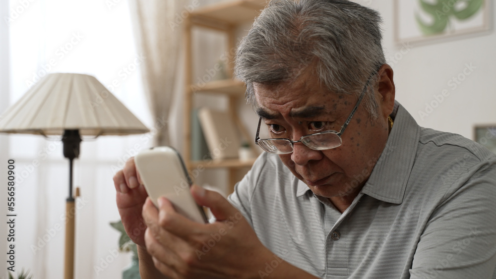 Elderly asian male having eyesight problems and wearing eyeglasses while gazing and reading message on smartphone on sofa. Old man having eye blurred vision indoors and moving glasses up and down Elderly asian male having eyesight problems and wearing eyeglasses while gazing and reading message on smartphone on sofa. Old man having eye blurred vision indoors and moving glasses up and down