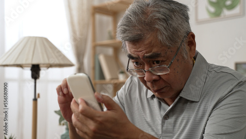 Elderly asian male having eyesight problems and wearing eyeglasses while gazing and reading message on smartphone on sofa. Old man having eye blurred vision indoors and moving glasses up and down