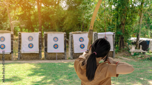 Back view of Asian girl wear face mask aims archery bow and arrow to colorful target in shooting range during training and competition. Exercise and concentration with outdoor archery.Archery for teen