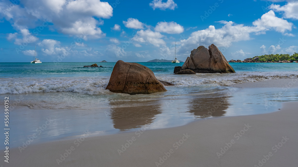 Picturesque granite rocks in the surf on a tropical beach. The waves of the turquoise ocean are foaming. Yachts and the outlines of islands on the horizon. Blue sky, clouds. Reflection on wet sand