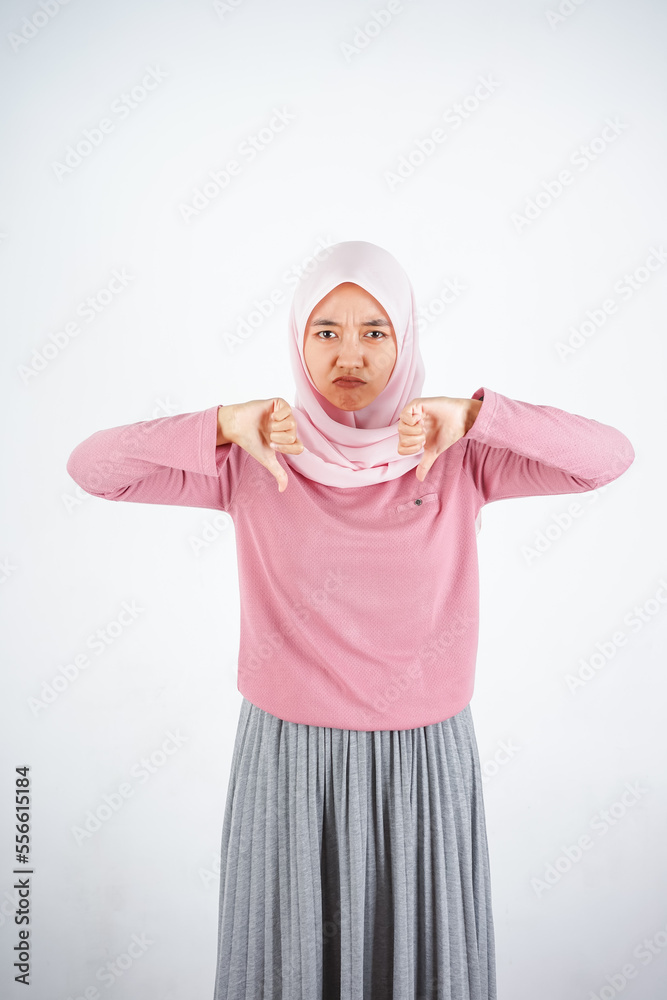 Beautiful Asian woman in pink sweater and hijab shows upset expression with thumbs down on white background Beautiful Asian woman in pink sweater and hijab shows upset expression with thumbs down on white background
