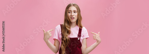 Self-assured arrogant young female student with high ego standing cocky and brag about herself pointing at her with cool snobbish look looking away to right with contempt posing over pink wall