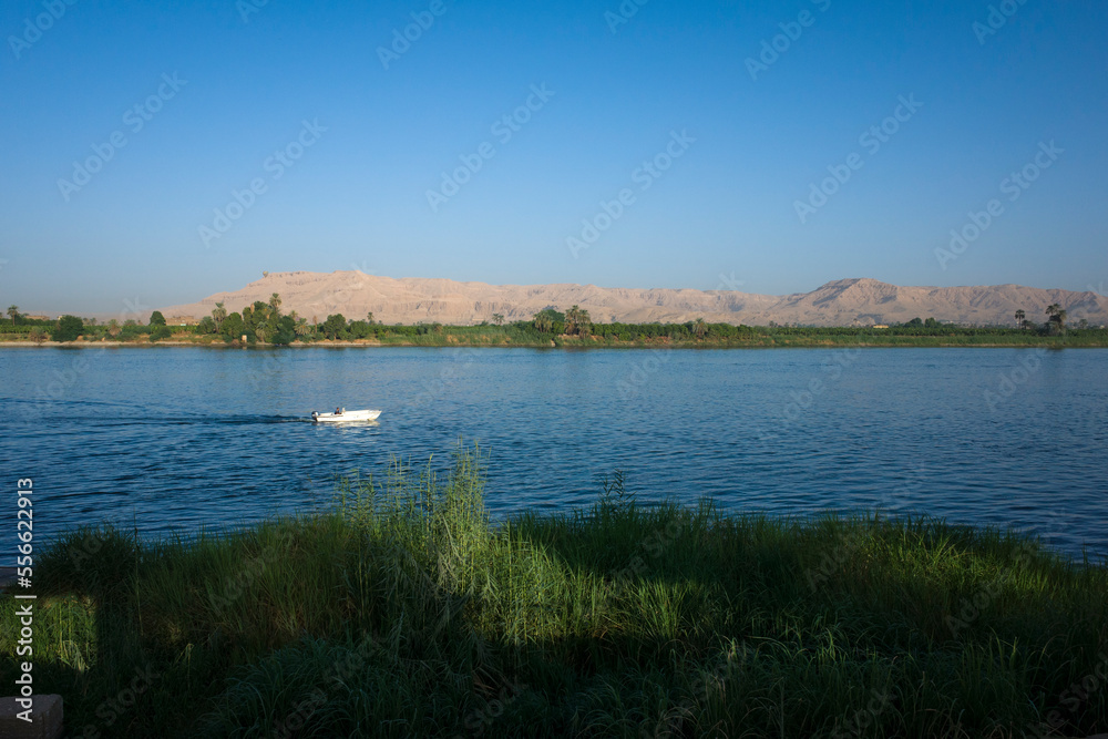 Nile river landscape in Luxor, Upper Egypt, White motorboat on blue water with view of Sahara desert dry mountains and green plantations on river west bank