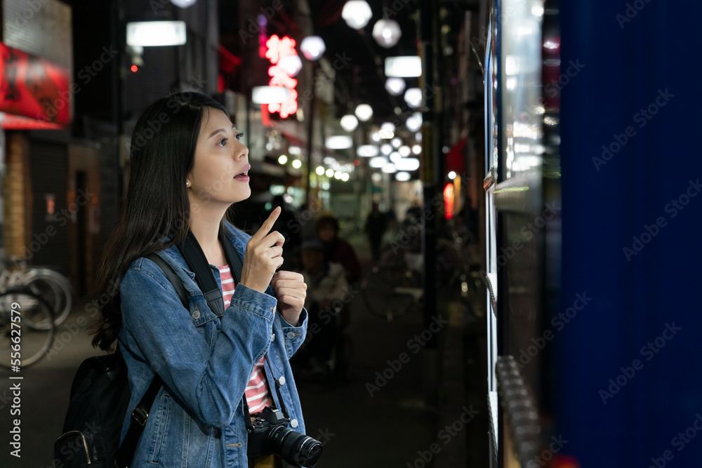 hesitant korean girl standing in front of vending machine thinking about which drink to choose on shopping street in Osaka japan at night. she looks at items with finger pointing