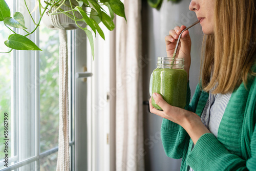 Woman drinking green smoothie standing by window at home photo