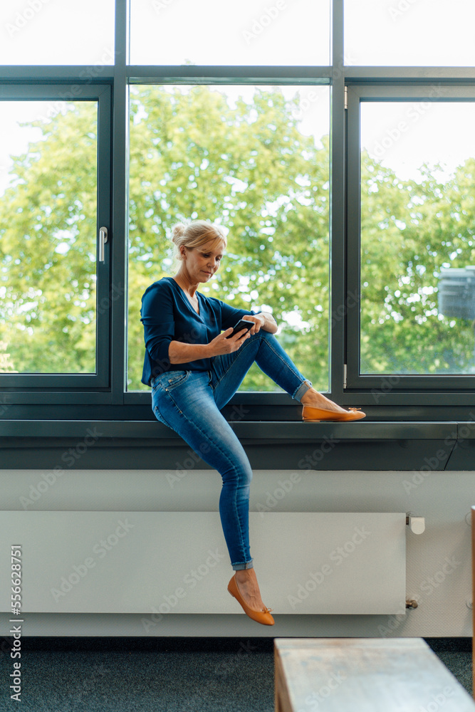 Businesswoman using smart phone sitting on window sill at office