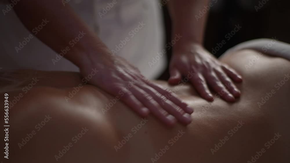 Close up cropped shot of unrecognizable male massage therapist gently stroke lower back of woman with beautiful skin and shiny from massage oil, lying on massage table in spa salon with dark interior. Close up cropped shot of unrecognizable male massage therapist gently stroke lower back of woman with beautiful skin and shiny from massage oil, lying on massage table in spa salon with dark interior.