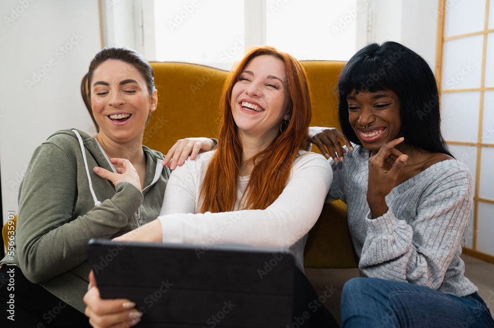 Group of three female friends with different ethnicity laughing and enjoying using tablet. Women cozy sitting on floor in living room. Group of three female friends with different ethnicity laughing and enjoying using tablet. Women cozy sitting on floor in living room.