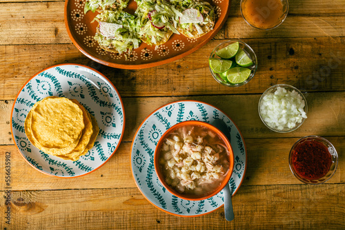 Pozole served in a deep dish, with tostadas, salsa and vegetables on a wooden table. Typical Mexican food.