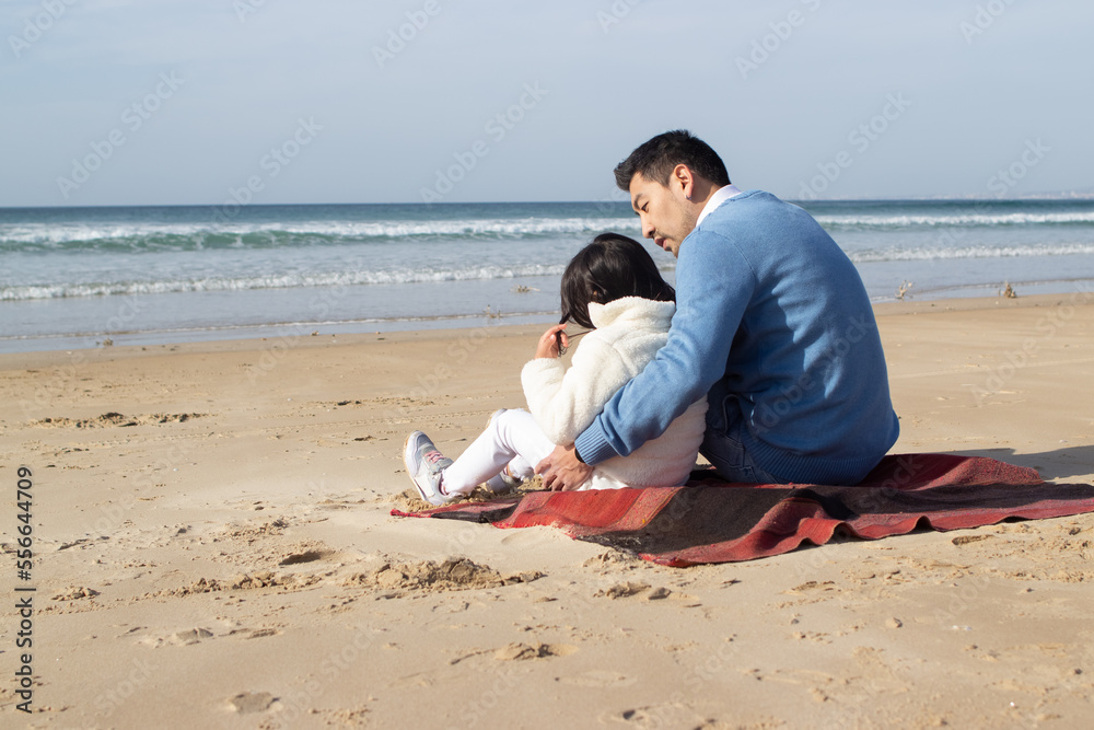 Happy Japanese family on blanket on beach. Father and daughter in casual clothes sitting on blanket, hugging. Family, relaxation, nature concept