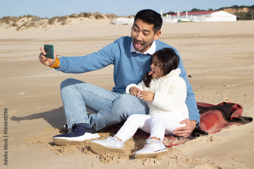 Merry Japanese father and daughter taking selfie at beach. Dark-haired child and man holding phone, making faces. Family, technology concept Merry Japanese father and daughter taking selfie at beach. Dark-haired child and man holding phone, making faces. Family, technology concept