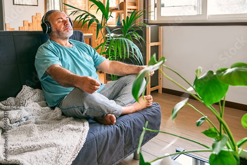Mature middle-aged overweight man in wireless headphones relaxing at home with guided meditation, listening to relaxing music on smartphone and meditating in lotus pose.