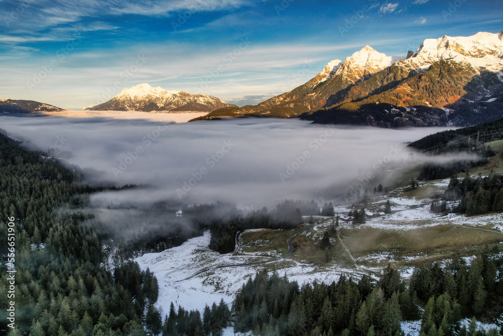 Dreamy mysterious winter landscape with fog and snow covered mountains and trees and blue bright sky with clouds