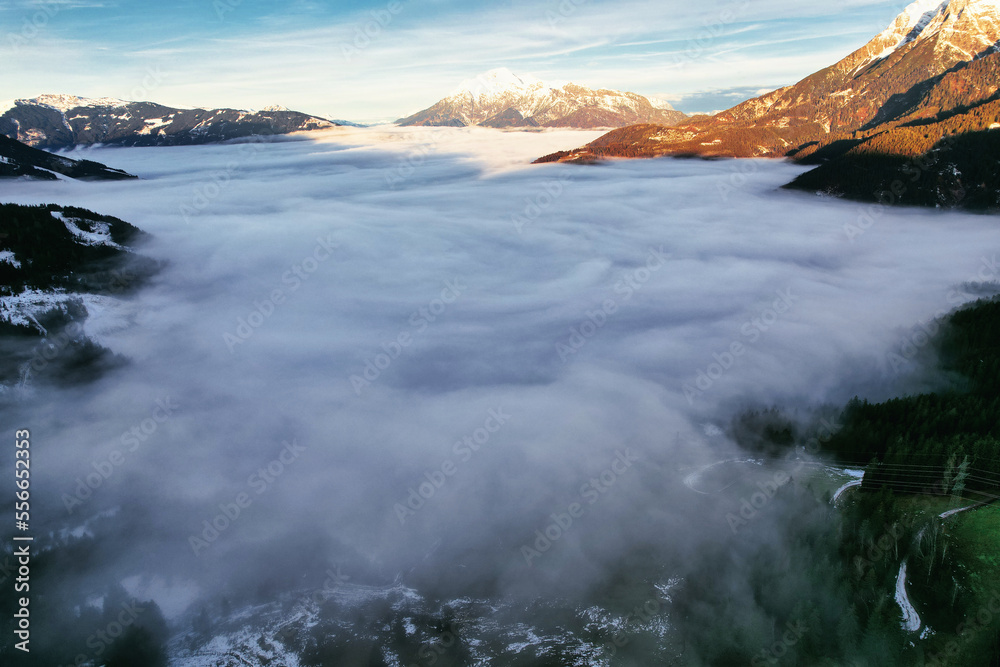 Dreamy mysterious winter landscape with fog and snow covered mountains and trees and blue bright sky with clouds