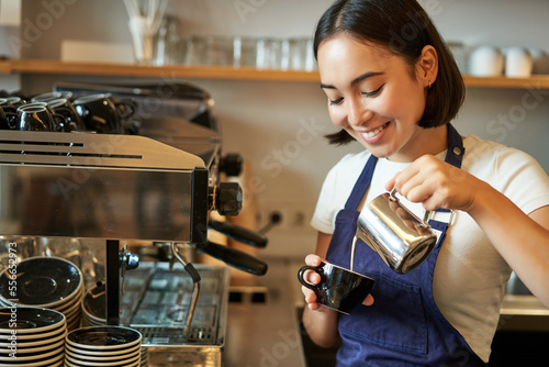 Close up of cute asian barista girl making cappuccino, doing latte art in cup with steamed milk, standing in coffee shop behind counter