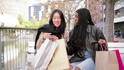 Two multiracial young women smiling and looking inside of a shopping bag sitting on a bench. A couple of happy friends showing their purchases on a sale week. Consumism concept. High quality photo photo