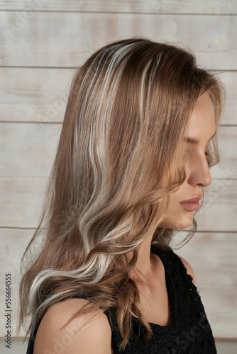 Close-up shot of a blonde girl with ash blonde wavy hair extensions. The girl in a black top with clip-in fake hair strands looks down on a light wooden background. Side view.