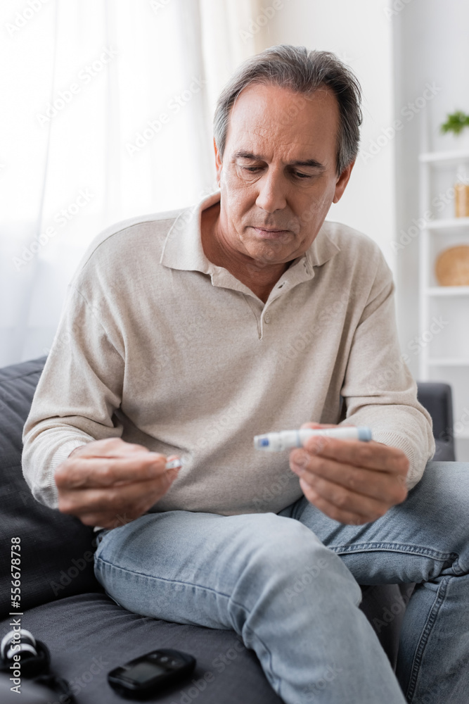 mature man with diabetes holding lancet pen and test strip while sitting on couch in living room