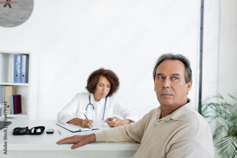 Mature man with diabetes sitting near blurred african american doctor in clinic Mature man with diabetes sitting near blurred african american doctor in clinic