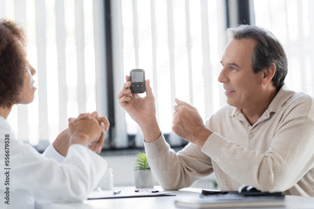Positive mature patient holding glucometer near blurred african american doctor in clinic Positive mature patient holding glucometer near blurred african american doctor in clinic