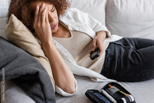 African american woman with diabetes touching head and holding glucometer on couch