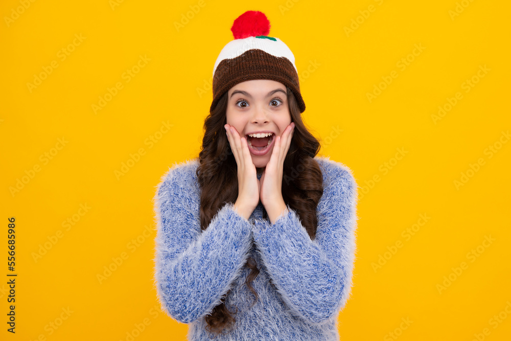 Teenager girl with winter hat over isolated yellow background. Winter christmas holidays, new year mood. Kids warm clothes. Shocked amazed face, surprised emotions of young teenager girl.