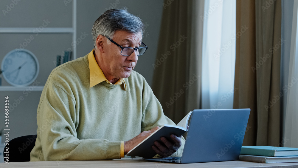 Senior older male online distant teacher webinar speaker tutor coach talking to webcam of laptop video training vlog online conference call old man holding notebook with notes reading information Senior older male online distant teacher webinar speaker tutor coach talking to webcam of laptop video training vlog online conference call old man holding notebook with notes reading information