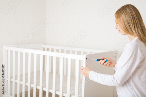 Fotografia Young adult woman using manual screwdriver and screwing white planks for newborn crib at nursery room