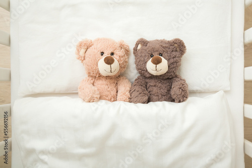 Two adorable smiling light and dark brown teddy bears lying down on pillow and sheet under blanket in baby crib. Twins concept. Top down view. Closeup.