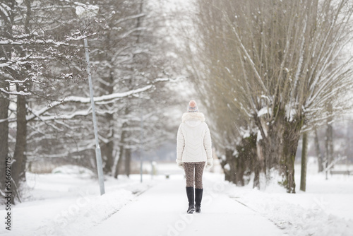 Young adult woman slowly walking on white snow covered sidewalk at park in cold winter day. Spending time alone. Enjoying peaceful stroll. Back view. © fotoduets