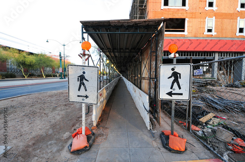 A covered pedestrian walkway runs alongside a construction site, flanked by directional signs and barriers, illustrating urban safety infrastructure and right-of-way accommodations during development