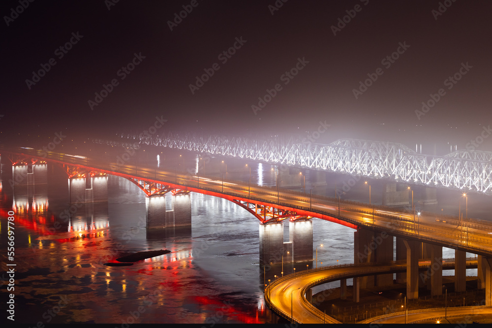 Night cityscape with two illuminated bridges across the Yenisei river disappearing into thick fog in Krasnoyarsk, Russia. Industrial smog in a city with bad ecology.Backlight road and railway bridges