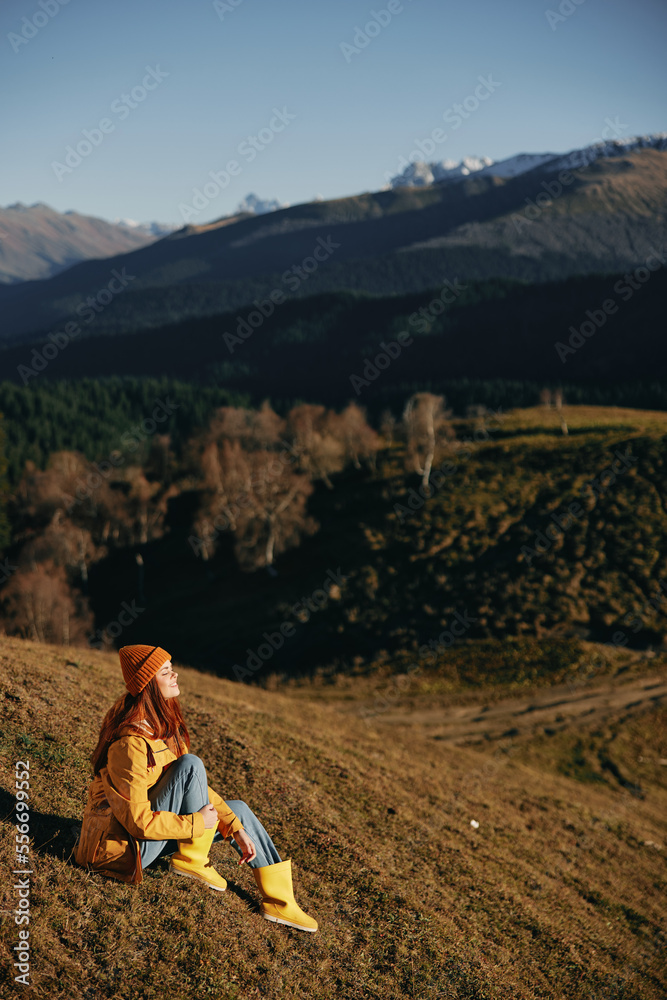 Fototapeta premium Woman full-length sitting resting on a hill smiling with teeth and looking at the mountains in a yellow raincoat and jeans happy camping trip in the spring, freedom lifestyle 