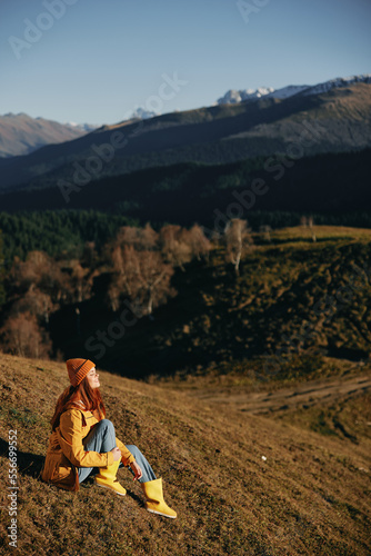 Woman full-length sitting resting on a hill smiling with teeth and looking at the mountains in a yellow raincoat and jeans happy camping trip in the spring, freedom lifestyle 