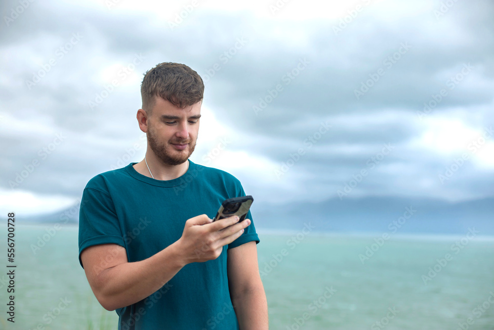 Happy guy, young handsome man is enjoying summer vacation at sea, beach in tropical exotic country, smiling, having fun, using his cell mobile phone, looking at smartphone