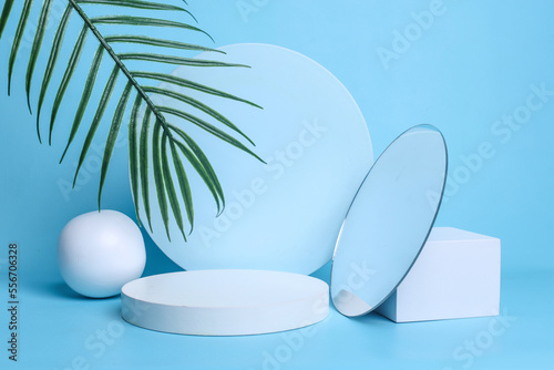 White circle podium with various geometric forms and palm leaves isolated on pastel blue background. Creative composition for beauty products presentation.