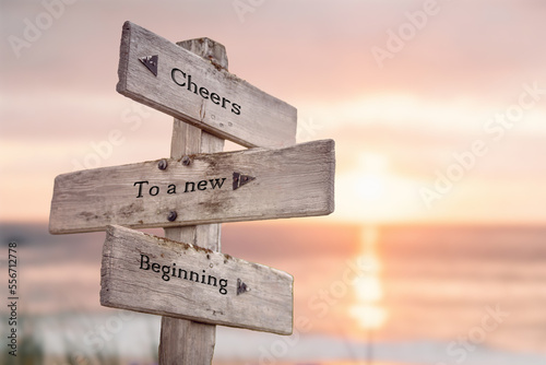 cheers to a new beginning text quote engraved on wooden signpost outdoors at the beach. Sunset theme.