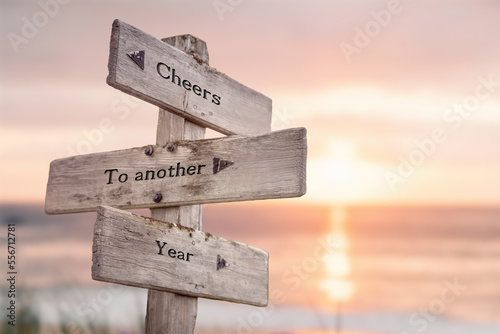 cheers to another year text quote engraved on wooden signpost outdoors at the beach. Sunset theme.