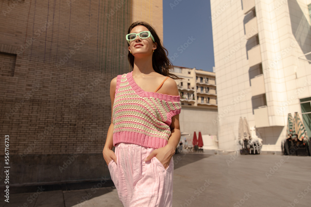 Bottom view of stylish young caucasian girl walks alone at european street in sunny day. Woman with brunette hair wears casual spring clothes. City life concept.
