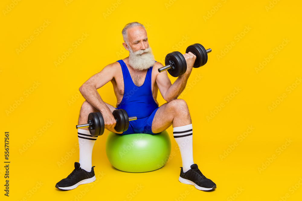 Full length photo of old senior grandfather gray beard hold hard weight dumbbel serious focused sitting ball biceps isolated on yellow color background Full length photo of old senior grandfather gray beard hold hard weight dumbbel serious focused sitting ball biceps isolated on yellow color background