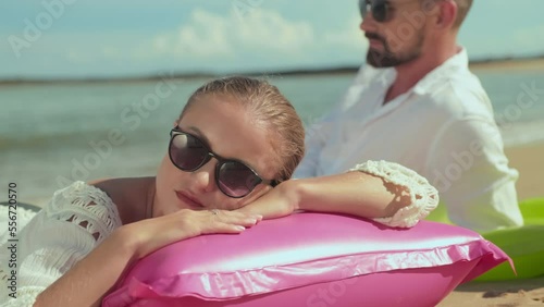 Young serene woman in sunglasses keeping head on pink inflatable mattress while sunbathing on tropical beach on hot sunny day against her husband relaxing on seashore