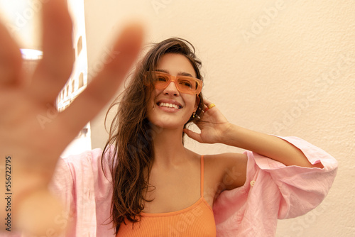Smiling young caucasian woman looking at distance streches hand at camera standing outdoor. Model with brunette wears yop, shirt and sunglasses. Concept of summer vacation. photo