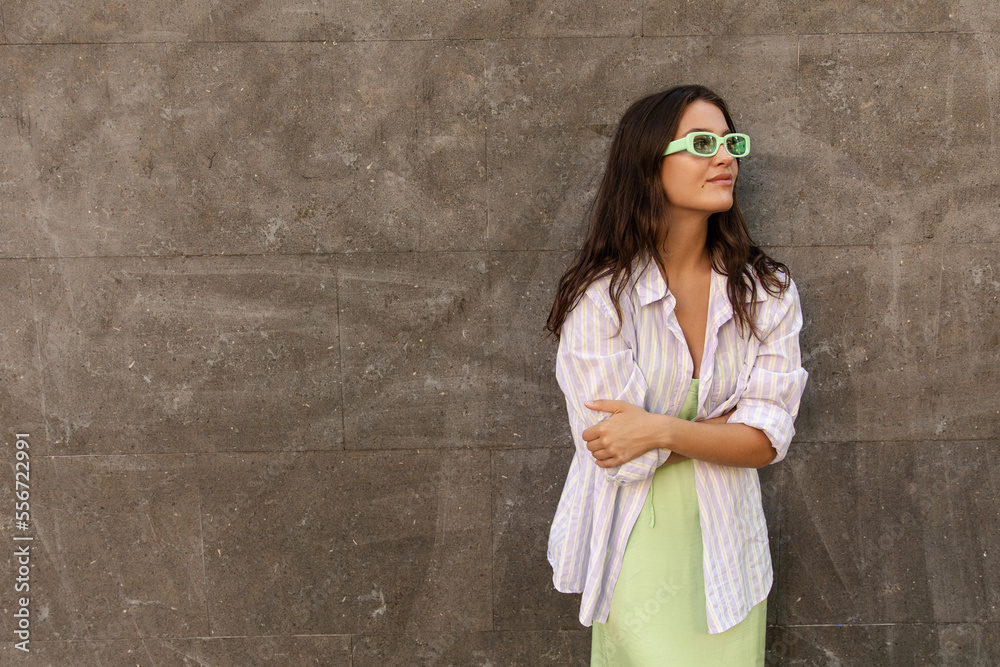 Calm young caucasian female looks away crossed her arms spends leisure spring time outdoor. Brunette wears shirt, sundress and sunglasses. Concept of city life, lifestyle.