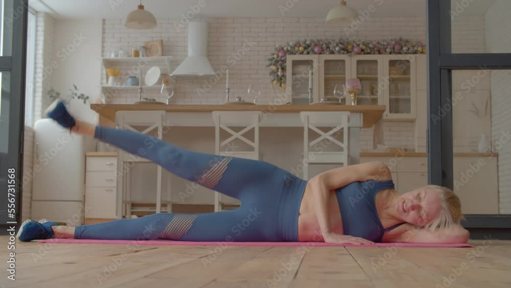 Active motivated charming sporty fitness senior female practicing side leg raises exercise on yoga mat, flexing abs, hip flexors, and lower back muscles improving core strength and stability at home.