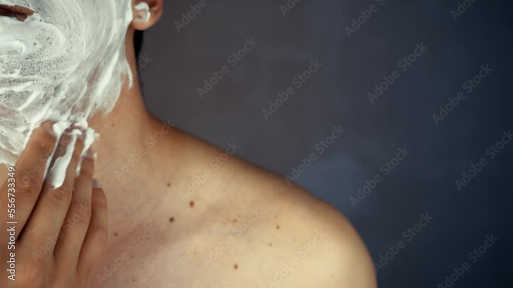 Handsome smiling unshaven young man applying moisturizing shaving foam on face and looking at camera. Portrait of happy man performing daily self-care, beauty routine indoors.