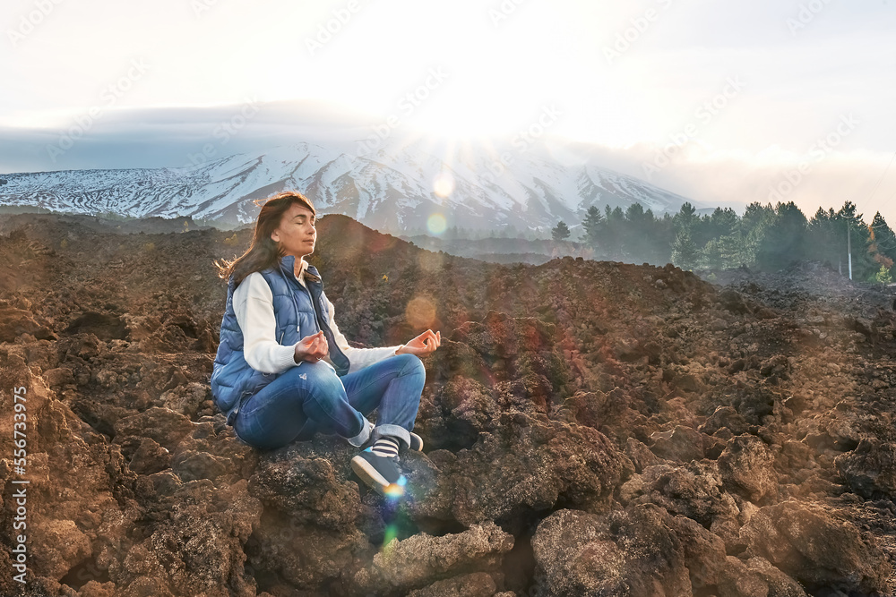 Happy tourist woman enjoying freedom while meditating on lava stone at panoramic view of snowy summits of active volcano Etna, Tallest volcano in Continental Europe, Sicily, Italy.