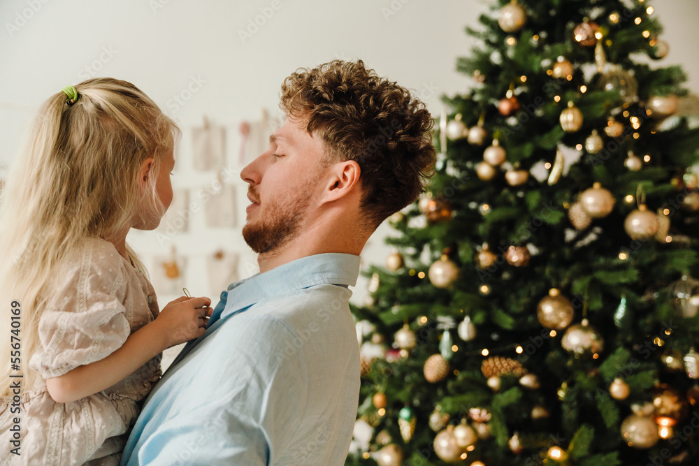 Father holding his daughter in his arms while standing near Christmas tree