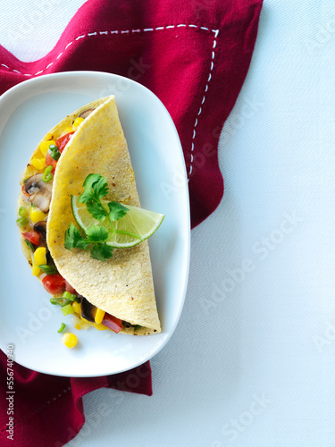 One vegetarian quesadilla in a yellow corn tortilla with cilantro, lime, mushrooms, corn, red pepper, melted cheese, and green onion. Mexican quesadilla is shown on a white cereamic plate with a red n photo